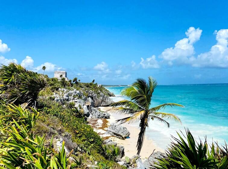 Side view of the Mayan ruins of Tulum right in front of the turquoise water and surrounded by palm trees.